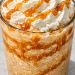 Tall glass of creamy caramel frappuccino topped with whipped cream and caramel drizzle, rich brown color with smooth texture.