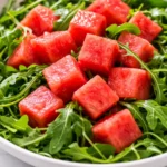 Fresh watermelon arugula salad with vibrant green leaves, juicy red watermelon cubes, and crumbled feta cheese, drizzled with balsamic glaze.
