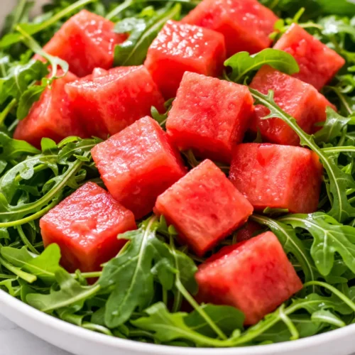 Fresh watermelon arugula salad with vibrant green leaves, juicy red watermelon cubes, and crumbled feta cheese, drizzled with balsamic glaze.