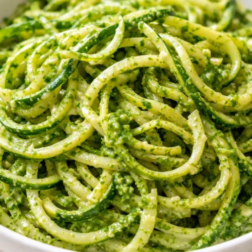 Creamy avocado pesto generously coats bright green spiralized zucchini noodles, garnished with cherry tomatoes and fresh basil leaves.