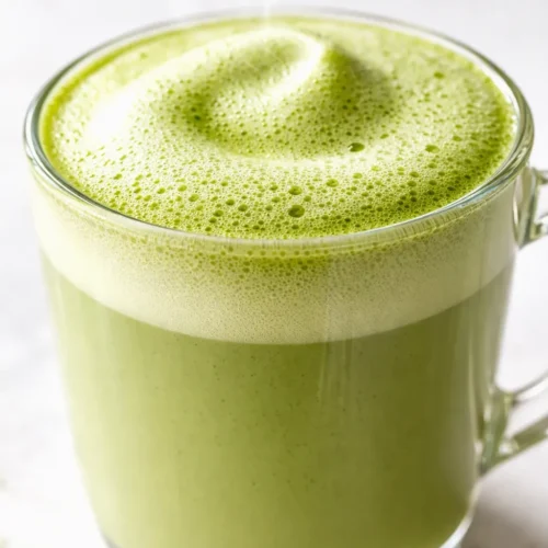 Creamy café-style matcha green tea latte in a tall glass showing vibrant green tea and frothy milk foam.