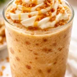 Creamy caramel crunch frappe in a tall glass, topped with whipped cream and caramel drizzle, garnished with crunchy toffee bits.