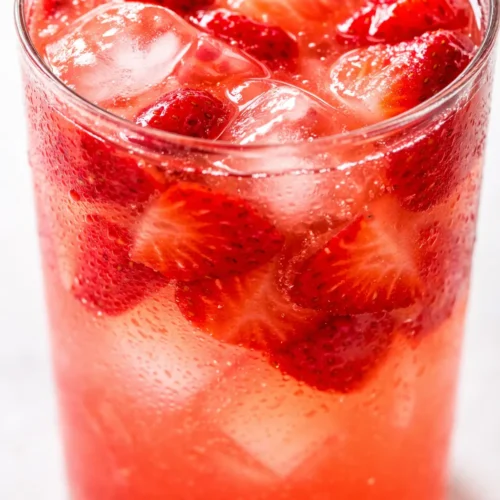 Bright pink strawberry lemonade in a glass pitcher with fresh strawberry slices, ice cubes, and mint leaves on a wooden table.