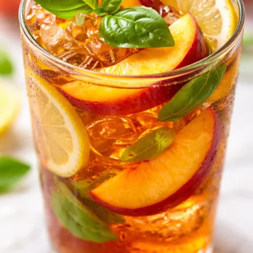 Golden amber iced tea with fresh peach slices and vibrant basil leaves over ice, a refreshing herbal summer drink.