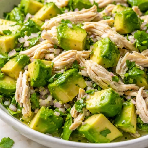 Creamy avocado chicken salad with fresh greens and cherry tomatoes in a white bowl, vibrant green and white chunks against a wooden table.