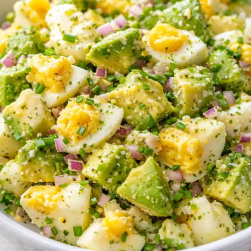 Creamy high protein avocado egg salad with chunky avocado pieces and sliced eggs, sprinkled with black pepper and fresh chives on a white plate.