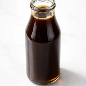 A wooden spoon stirs dark amber homemade chai syrup in a stainless steel pot, with cinnamon sticks and star anise visible, thick and glossy.