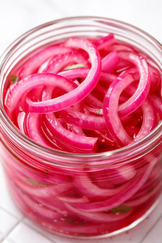 Vibrant magenta pickled red onion slices in a glass jar, crisp and glossy, showcasing their crunchy texture after a quick 15-minute prep.