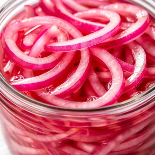 Vibrant magenta pickled red onions in a glass jar, showing crisp translucent slices submerged in tangy brine.