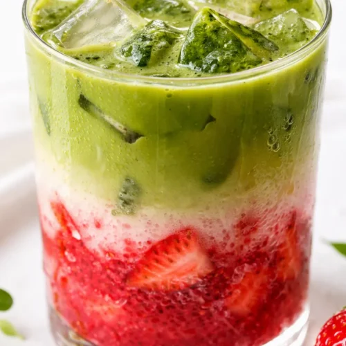 Iced strawberry matcha latte in a tall glass with layered red strawberry milk and green matcha, garnished with berries and ice.