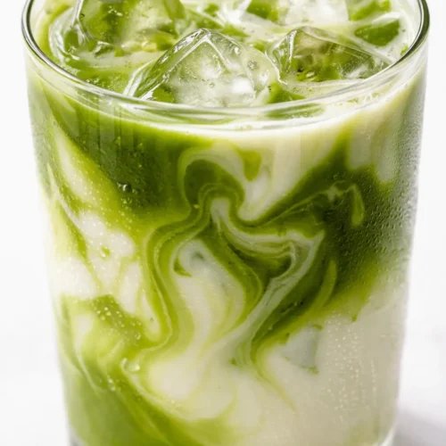 Creamy iced vanilla matcha latte with layered milk, vibrant green matcha, and vanilla syrup swirling over ice in a tall glass.