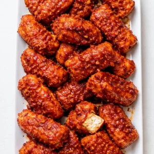 Baked buffalo tofu wings with golden crispy edges coated in glossy red hot sauce, resting on a parchment-lined baking sheet.