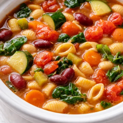 Hearty slow cooker minestrone in a rustic bowl, chunky vegetables and tender beans in rich red broth, topped with fresh basil and Parmesan.