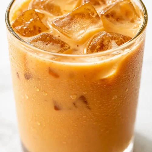 Tall glass of iced pumpkin spice latte with whipped cream, pumpkin drizzle, and cinnamon dusting, showing creamy orange layers.
